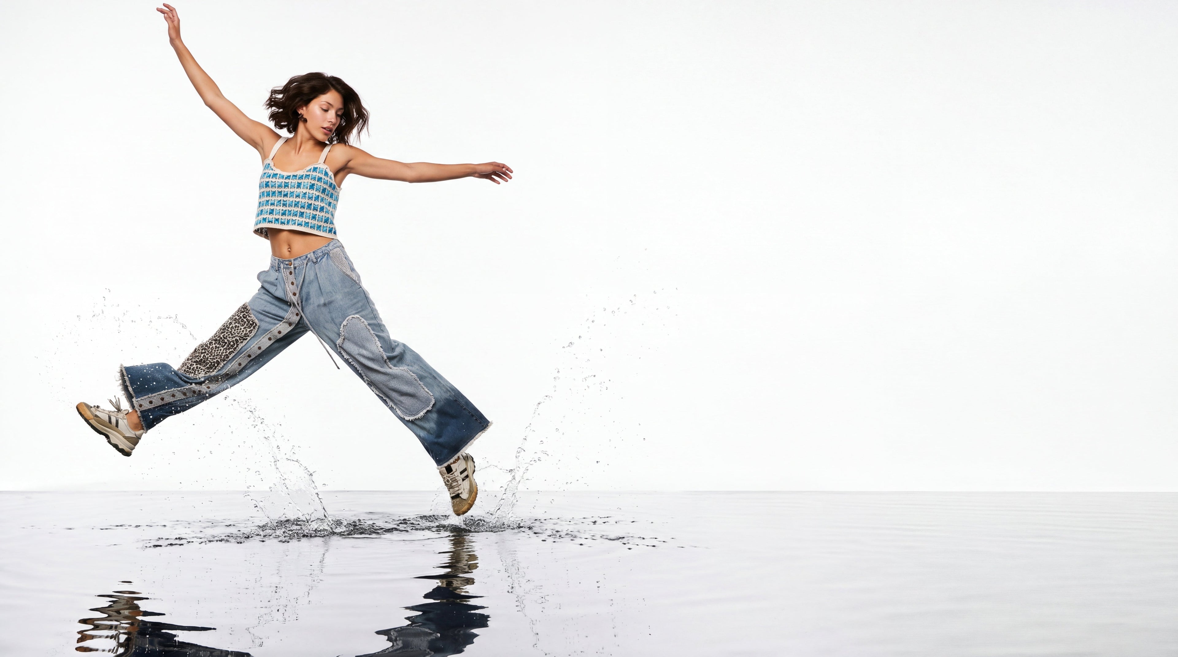 A woman jumps across a reflective body of water wearing a blue patterned crop top, wide-leg denim jeans with patterned side panels, and sneakers, against a bright white sky.